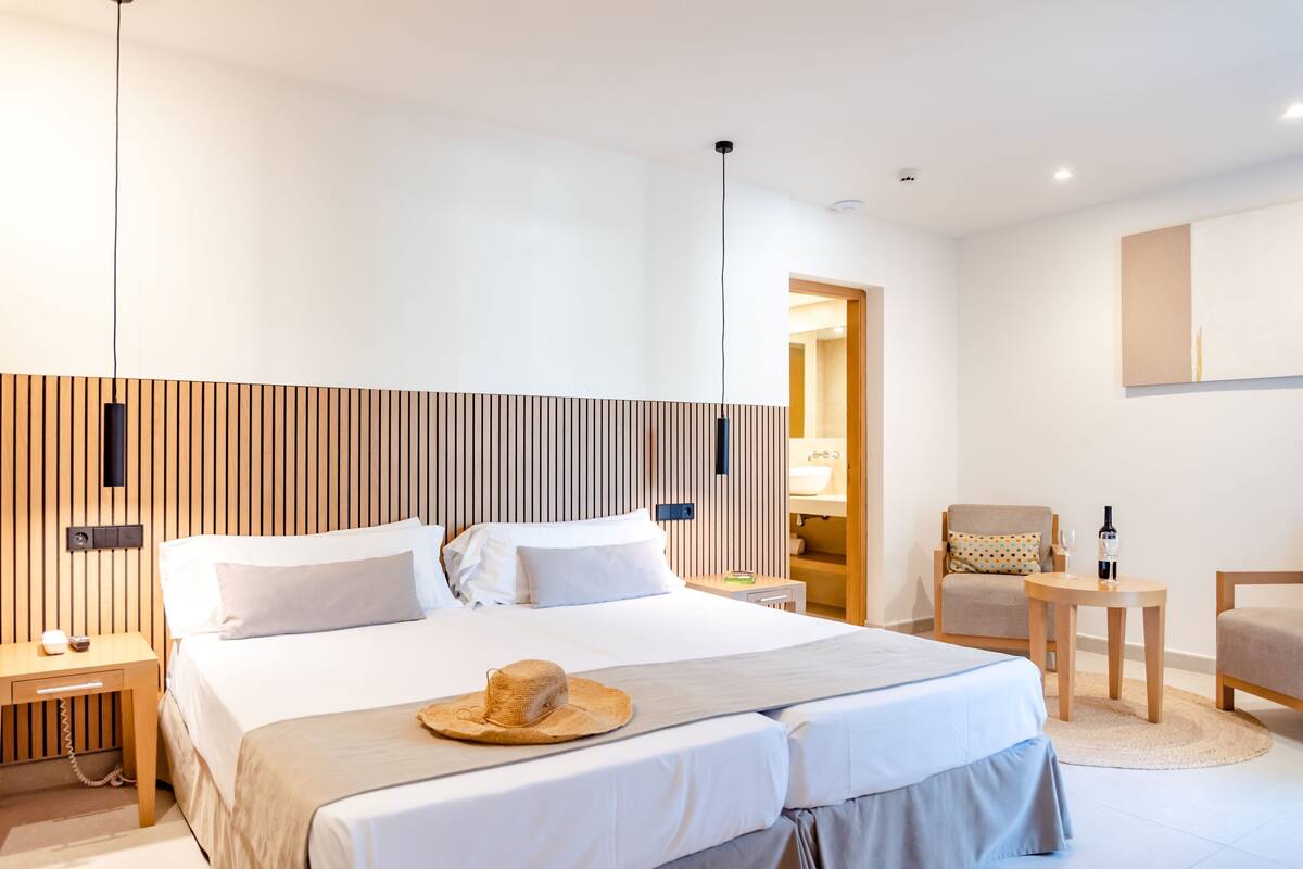 A modern hotel room with a large bed, a wooden headboard, a bedside table with a lamp, a chair, and a small table with a bottle and glass.