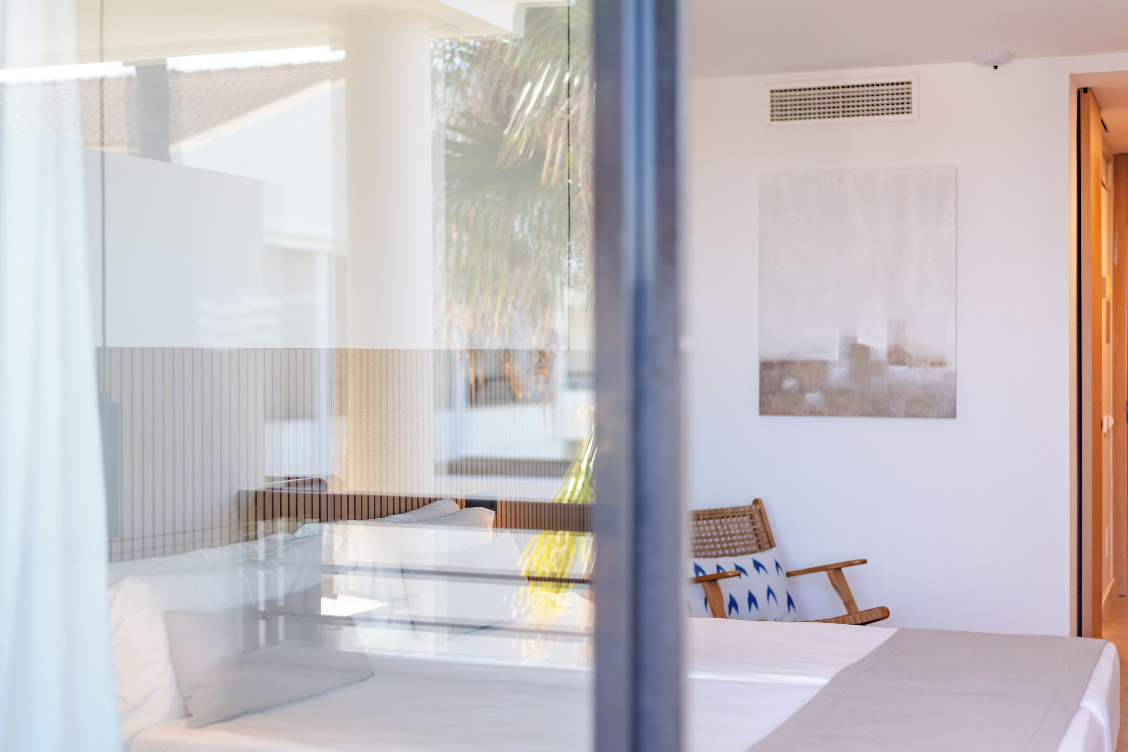 Una habitación de hotel moderna con una cama, una pequeña zona de estar con una silla y cojines, un ventanal grande y una obra de arte colgada en la pared.