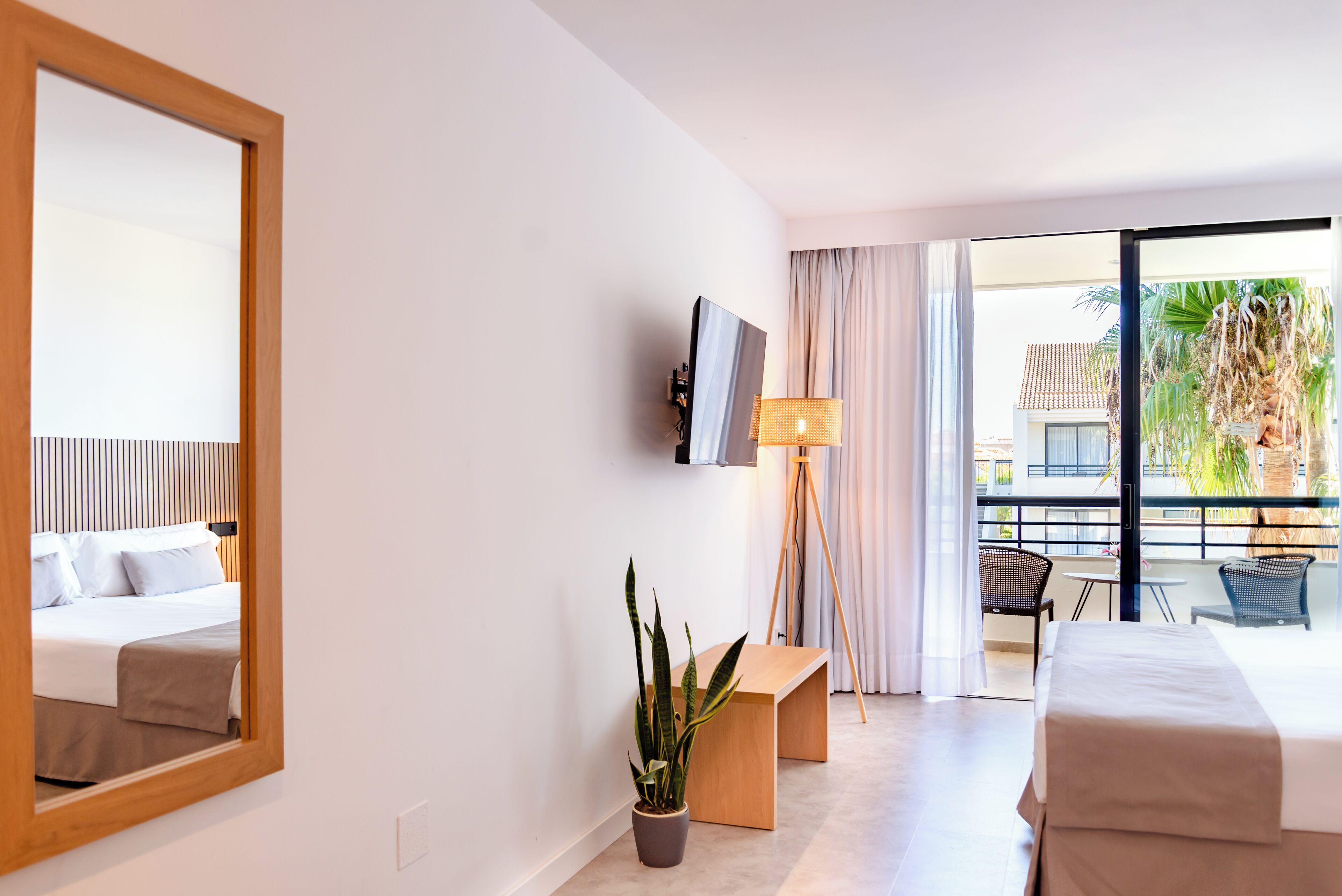 Habitación de hotel con un espejo grande, una cama, una mesita de noche, una planta en maceta, un televisor y un balcón con vistas a un edificio y palmeras.