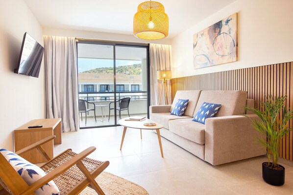 A modern living room with a sofa, a lounge chair, a small table, a TV, and a balcony with a view. - Hotel Eu Bahia Pollença (Pollenca)