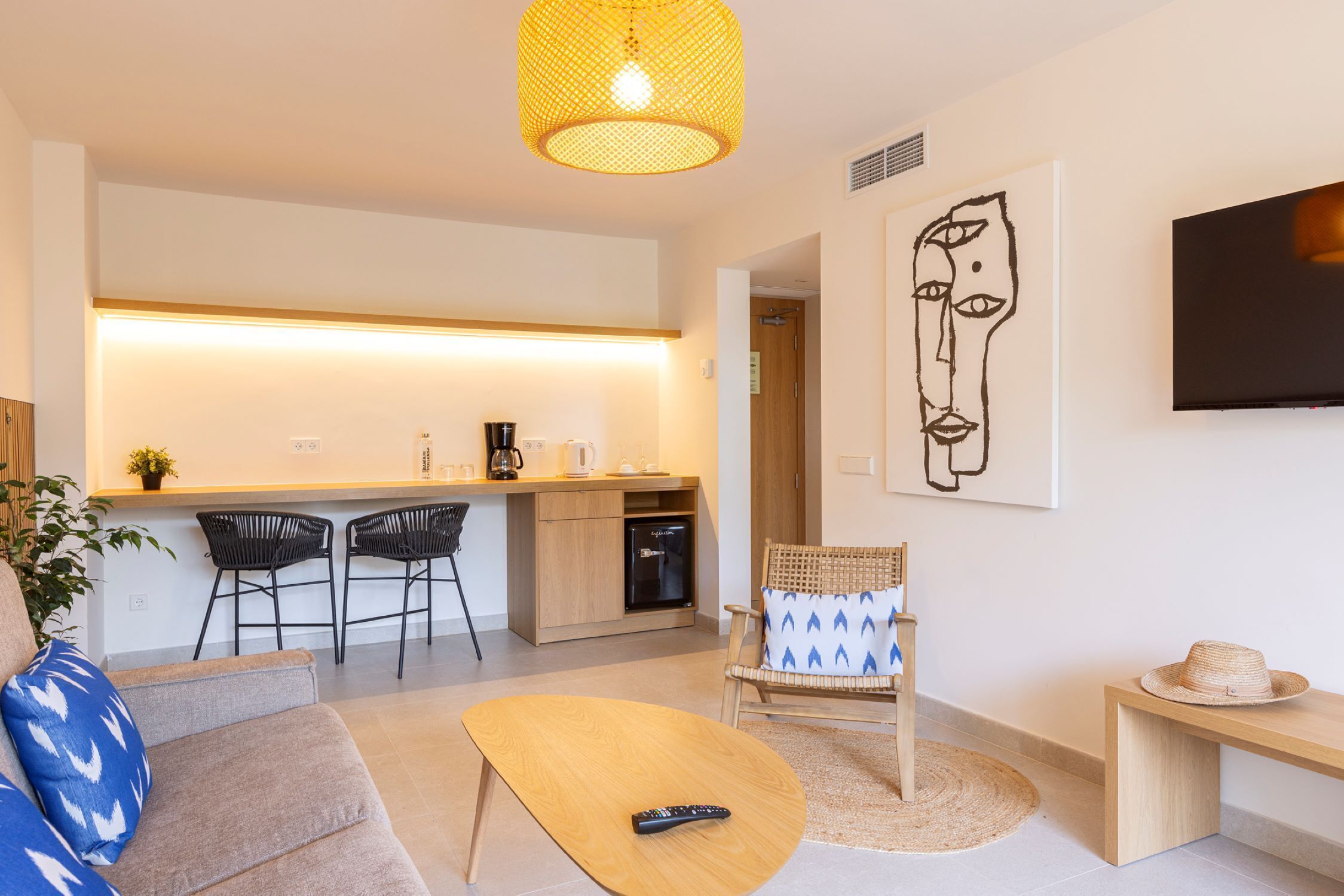 A modern living room with a sofa, coffee table, and a chair. There is a kitchen area with a counter and chairs. A television is mounted on the wall.