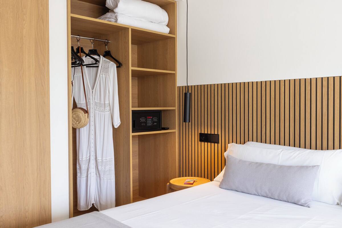 A hotel room with a bed, a wooden wardrobe, a small bedside table, and a wall-mounted control panel.
