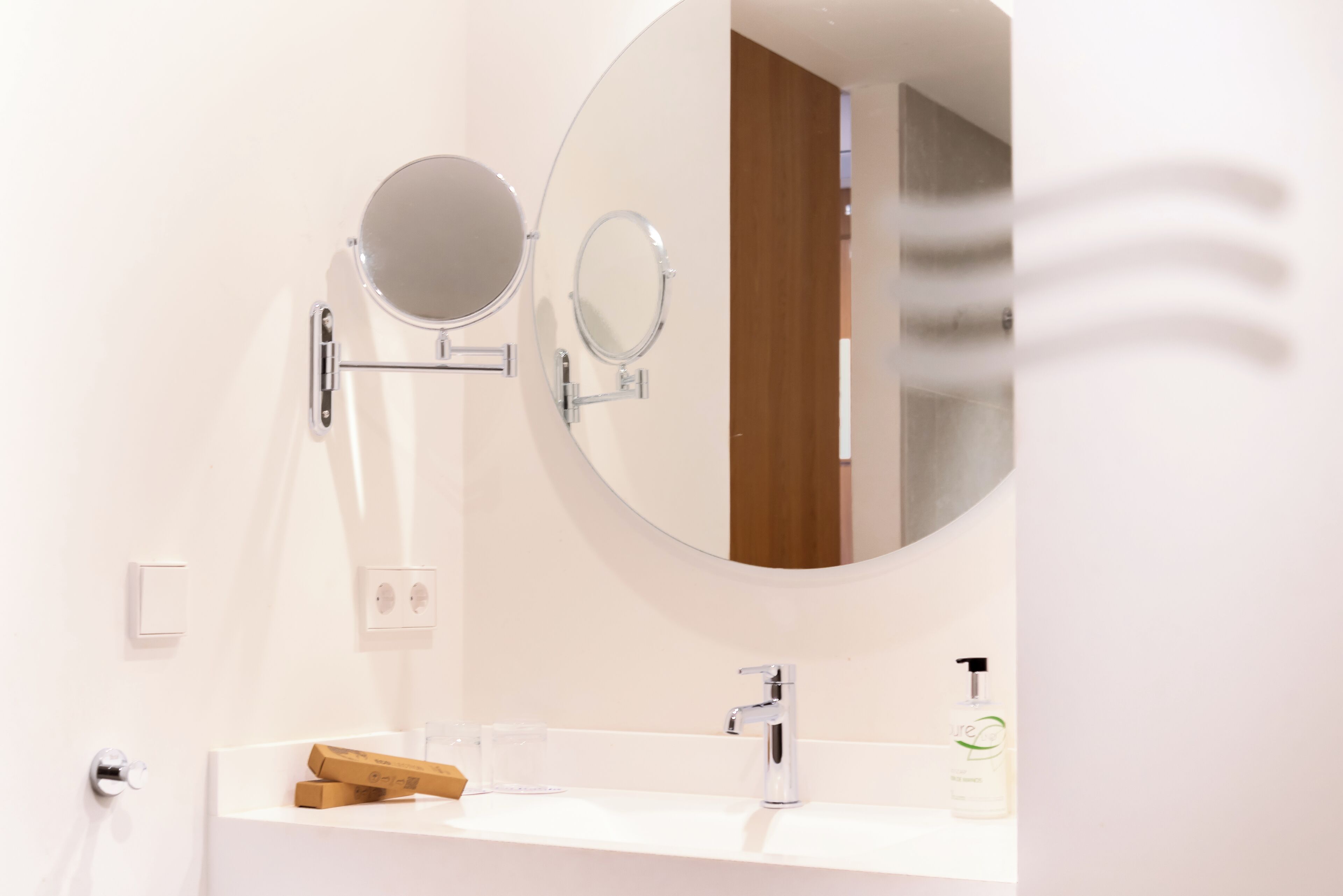 Une salle de bain avec un lavabo blanc, deux miroirs ronds, une douchette à main et un distributeur de savon.