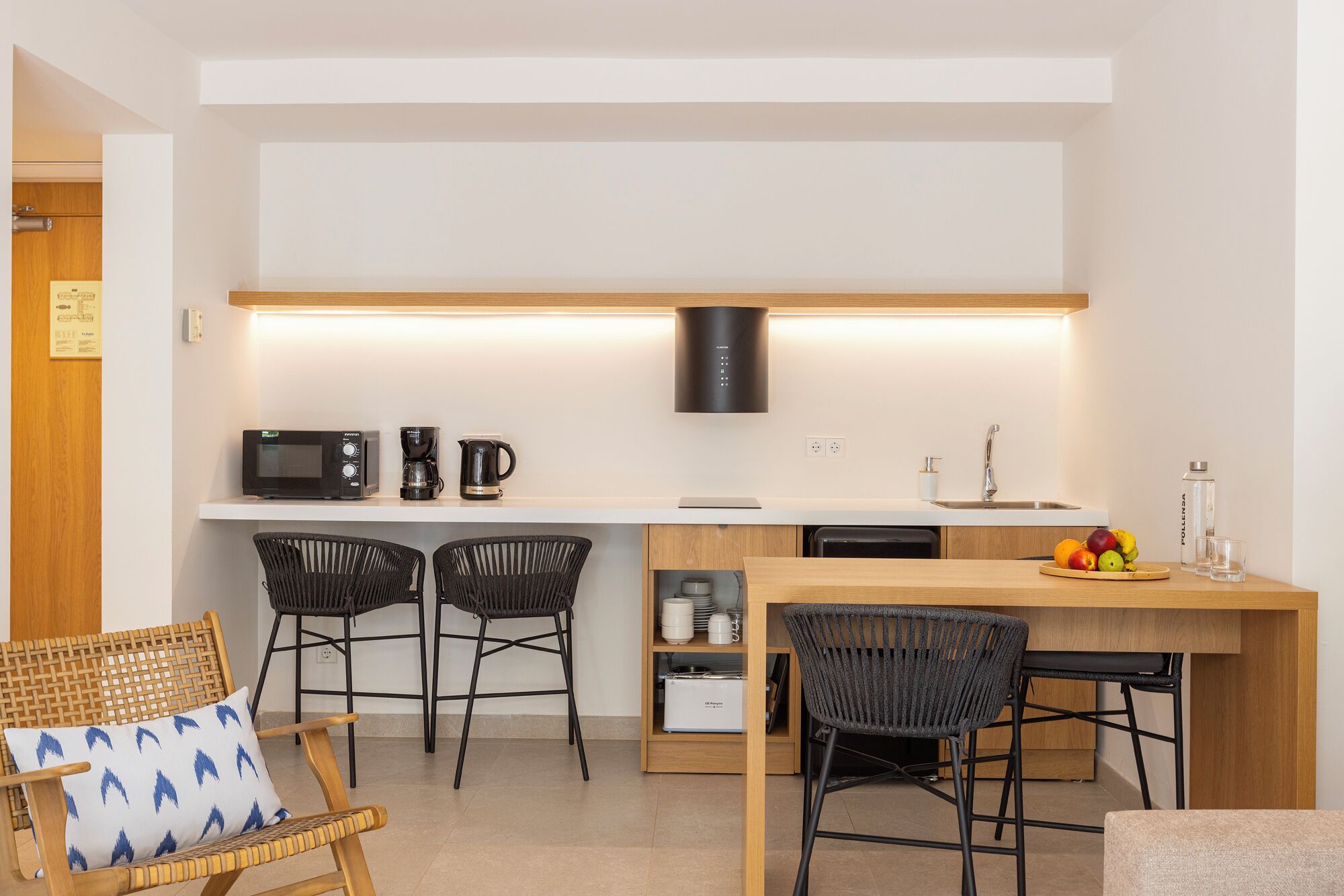 A modern kitchen with a microwave, coffee maker, and kettle on a white countertop. There are two black chairs and a wicker chair with a blue pillow. A wooden dining table with fruit and glasses is also visible.