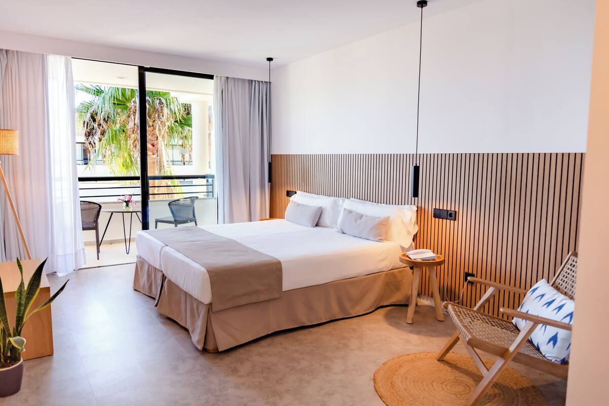 A modern hotel room with a large bed, a small side table, a chair, and a balcony with a view of palm trees.