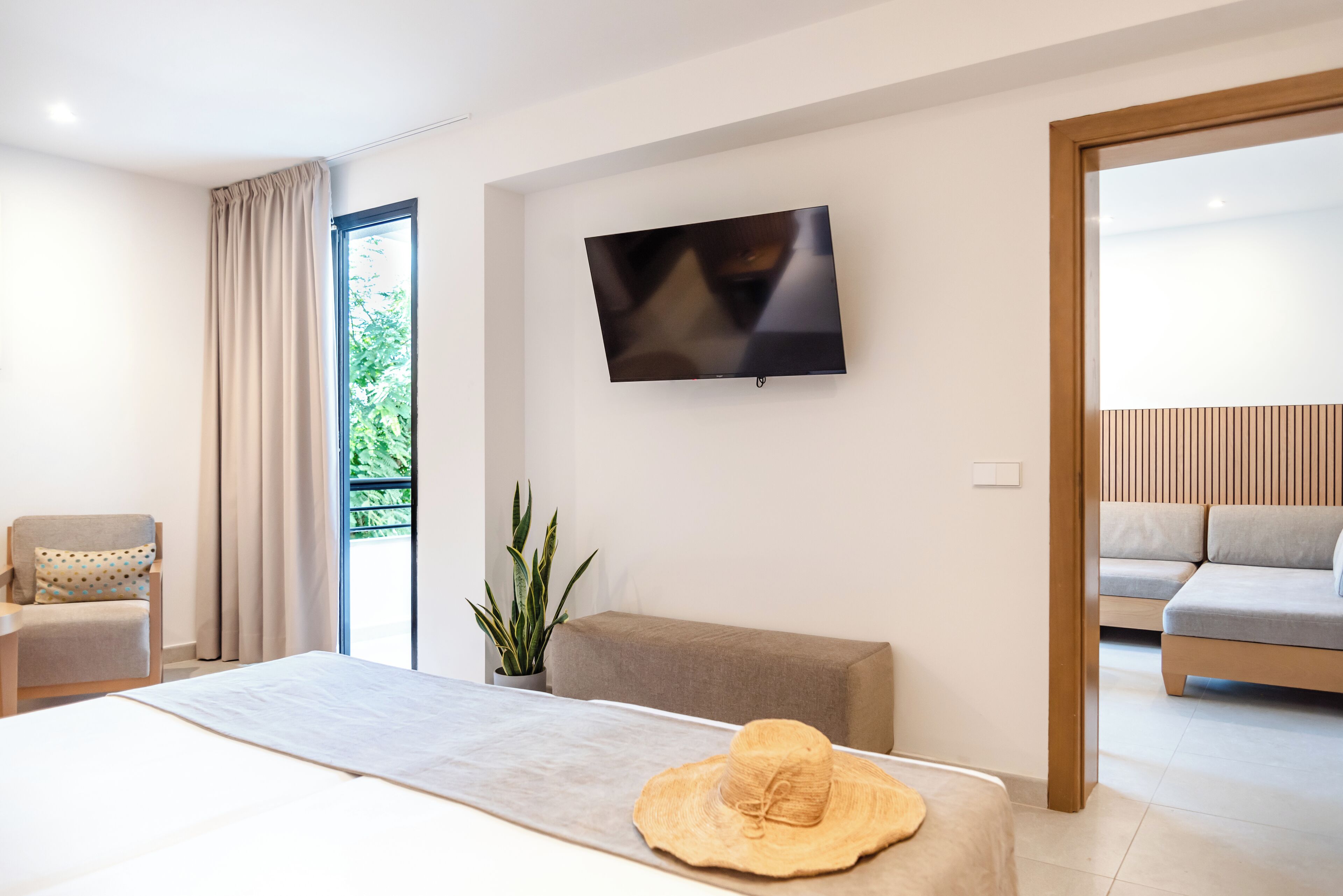 Een moderne hotelkamer met een bed, een aan de muur bevestigde televisie, een zithoek met een sofa en uitzicht op groen buiten.