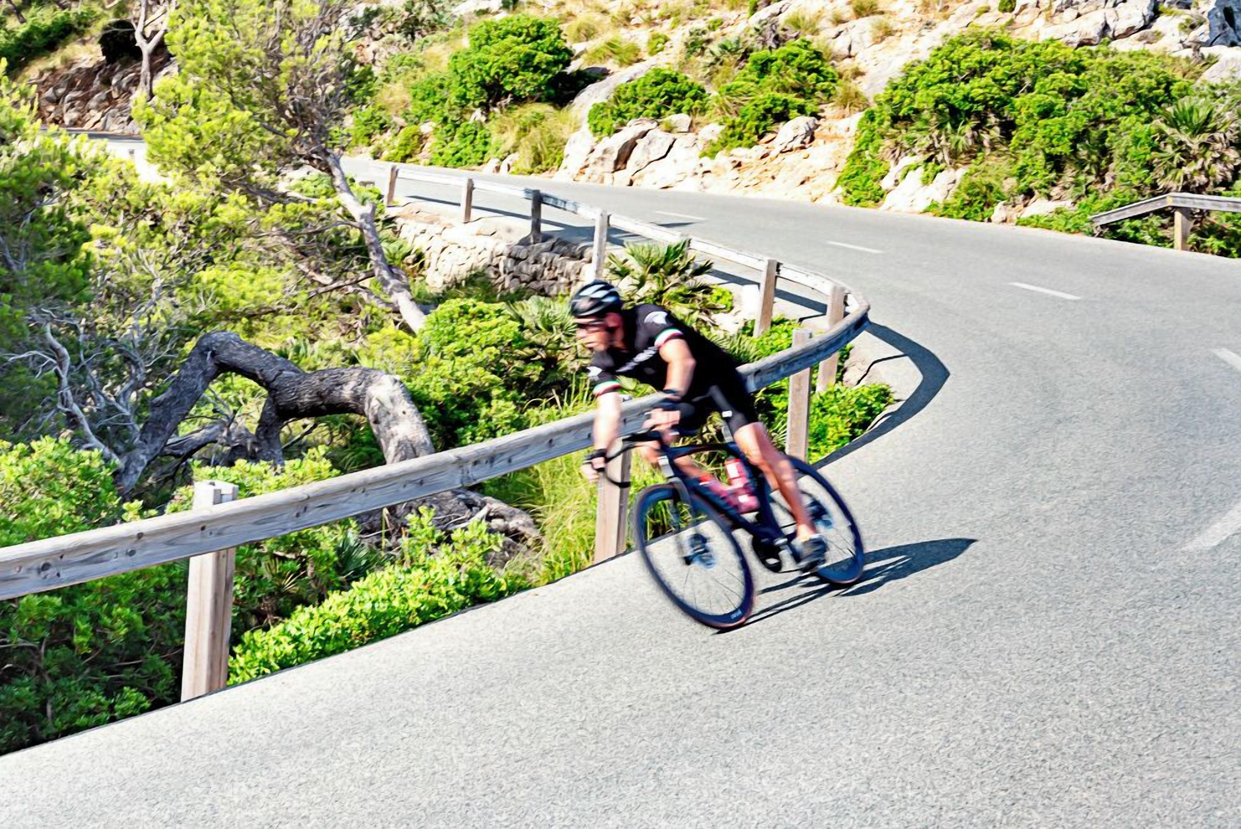 A cyclist riding on a paved road with a guardrail, surrounded by greenery and rocky terrain.