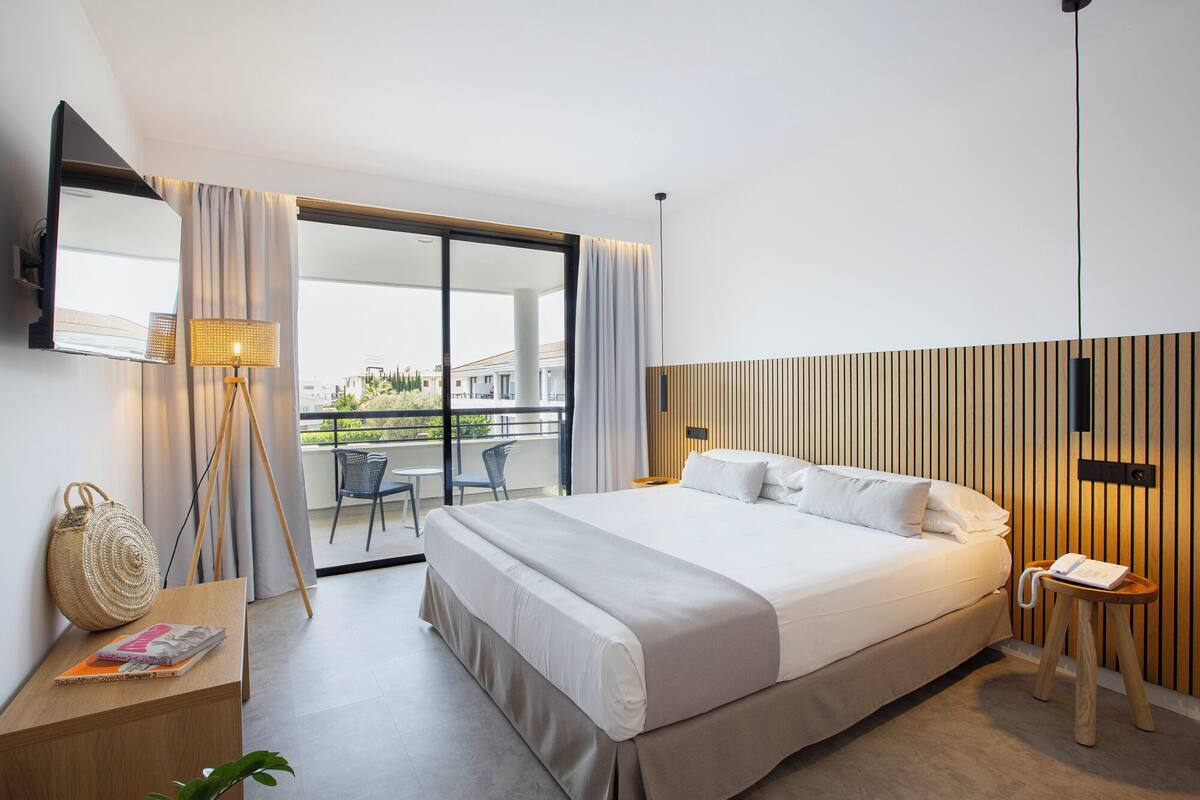 A modern hotel room with a large bed, a desk with a lamp, a small table with a phone, and a balcony with chairs.
