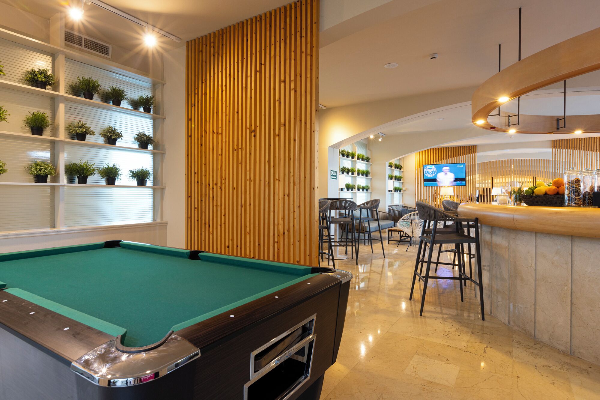 Una mesa de billar en un espacio interior bien iluminado, con estanterías llenas de plantas, una zona de bar con asientos y una pantalla de televisión.