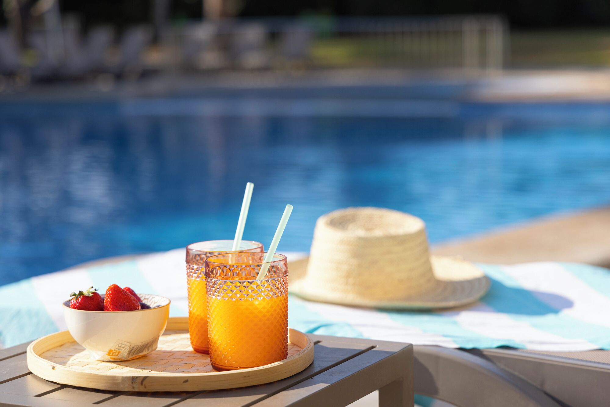 Escena junto a la piscina con una mesa que sostiene un tazón de fresas, dos vasos de jugo de naranja y una pajita. También se ve un sombrero de paja sobre la mesa.