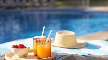 Escena junto a la piscina con una mesa que sostiene un tazón de fresas, dos vasos de jugo de naranja y una pajita. También se ve un sombrero de paja sobre la mesa.