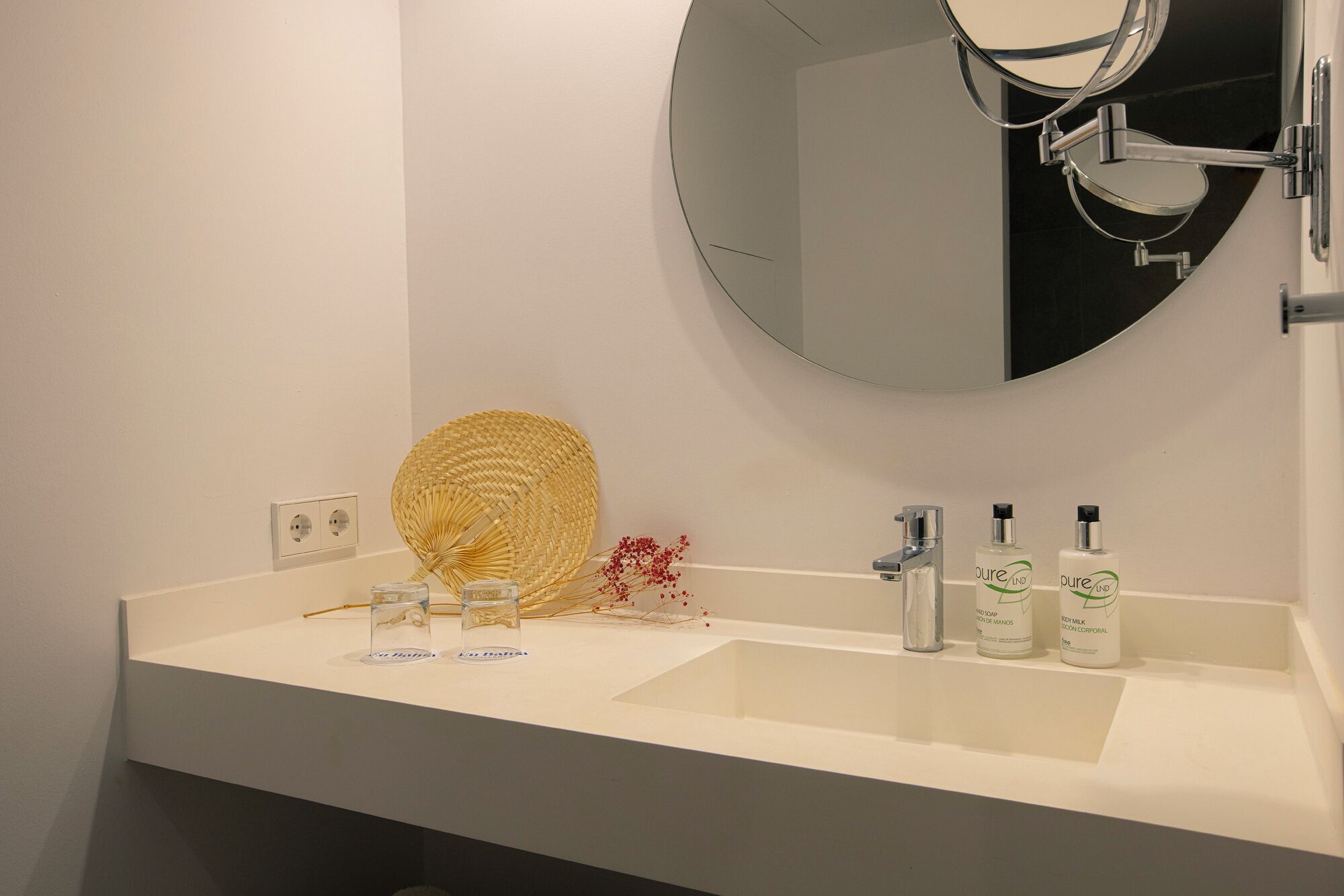 Un baño con un lavamanos blanco, un espejo redondo y tres botellas de jabón corporal.