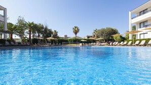 A clear blue pool with lounge chairs and umbrellas, surrounded by palm trees and a building with balconies.