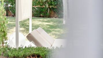 A white outdoor seating area with a cushioned chair and a white table, surrounded by greenery and a white structure.
