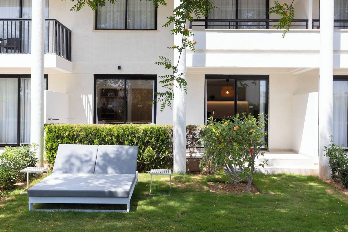 A modern building with a white exterior, large windows, and a well-maintained garden with a bench and small trees.