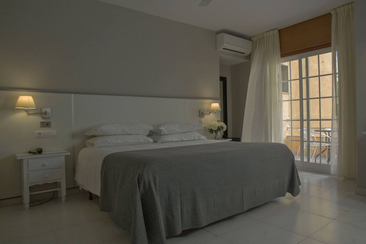 A hotel room with a bed, a nightstand, a lamp, a vase with flowers, a chair, and a window with curtains.