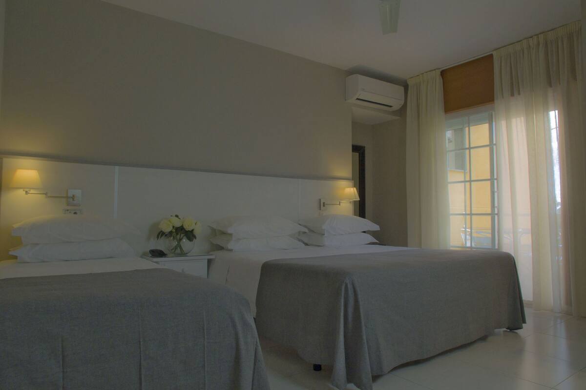 A hotel room with two beds, a nightstand with a lamp, an air conditioning unit, and a window with curtains.