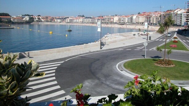 A coastal road with a pedestrian crossing, a roundabout, and a view of the sea and buildings in the background. - Hotel Ancora (Sanxenxo)