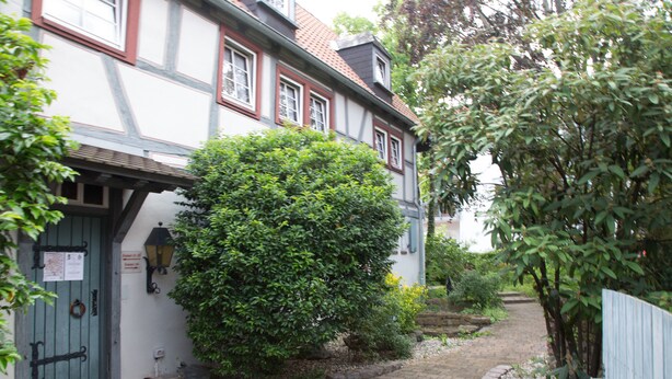 Un edificio tradicional con entramado de madera y un camino de piedra que conduce a una puerta verde, rodeado de abundante vegetación.