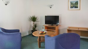 Una habitación con televisión, dos sillas azules, una mesa de madera con fruta, una planta en maceta y un cuadro en la pared.