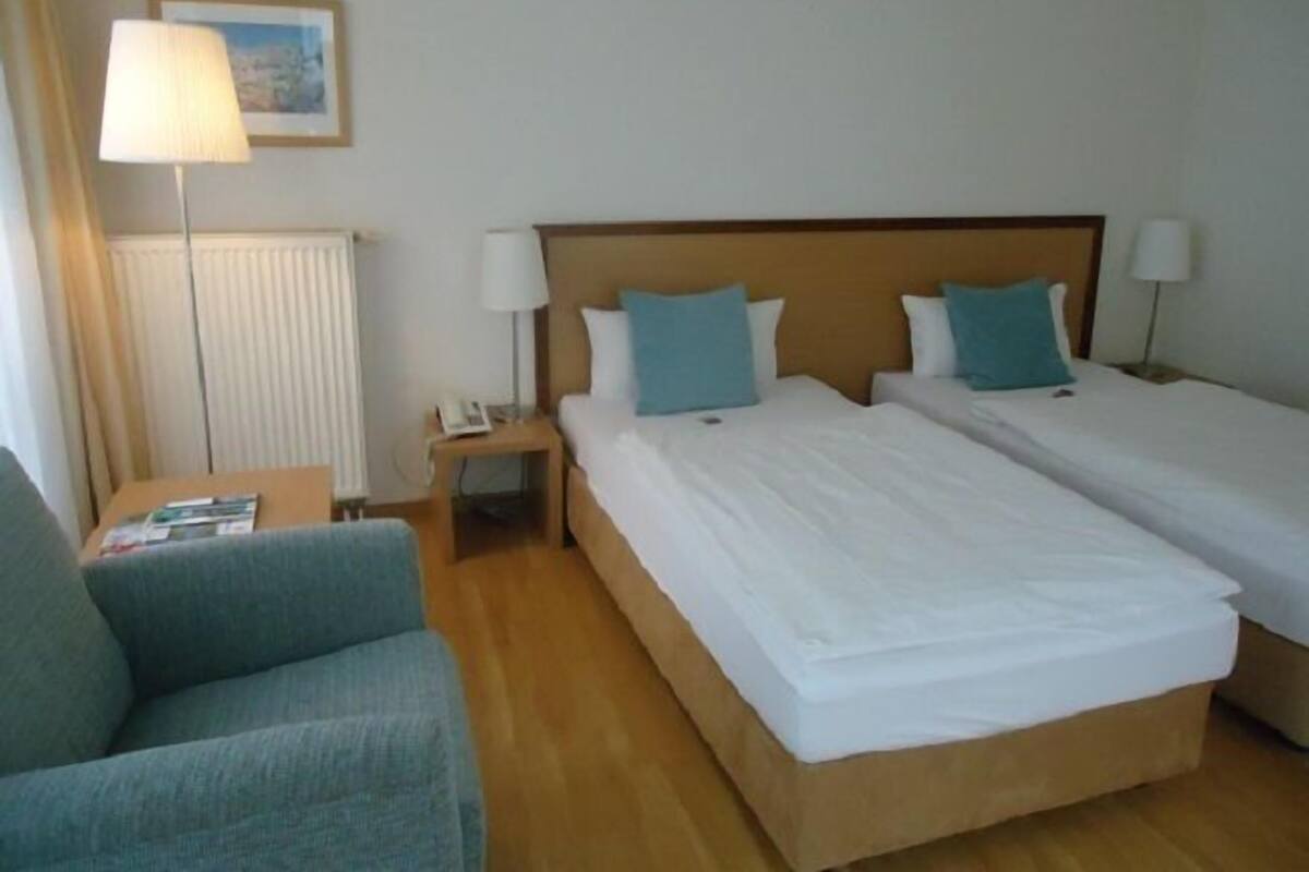 A hotel room with two single beds, a small table, a chair, and a lamp.