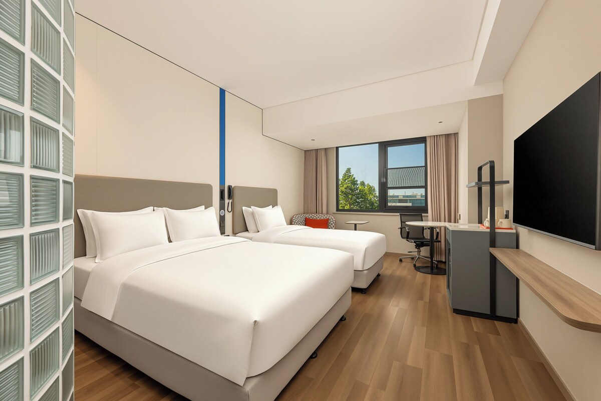 A hotel room with two beds, a desk, a TV, and a window with a view.