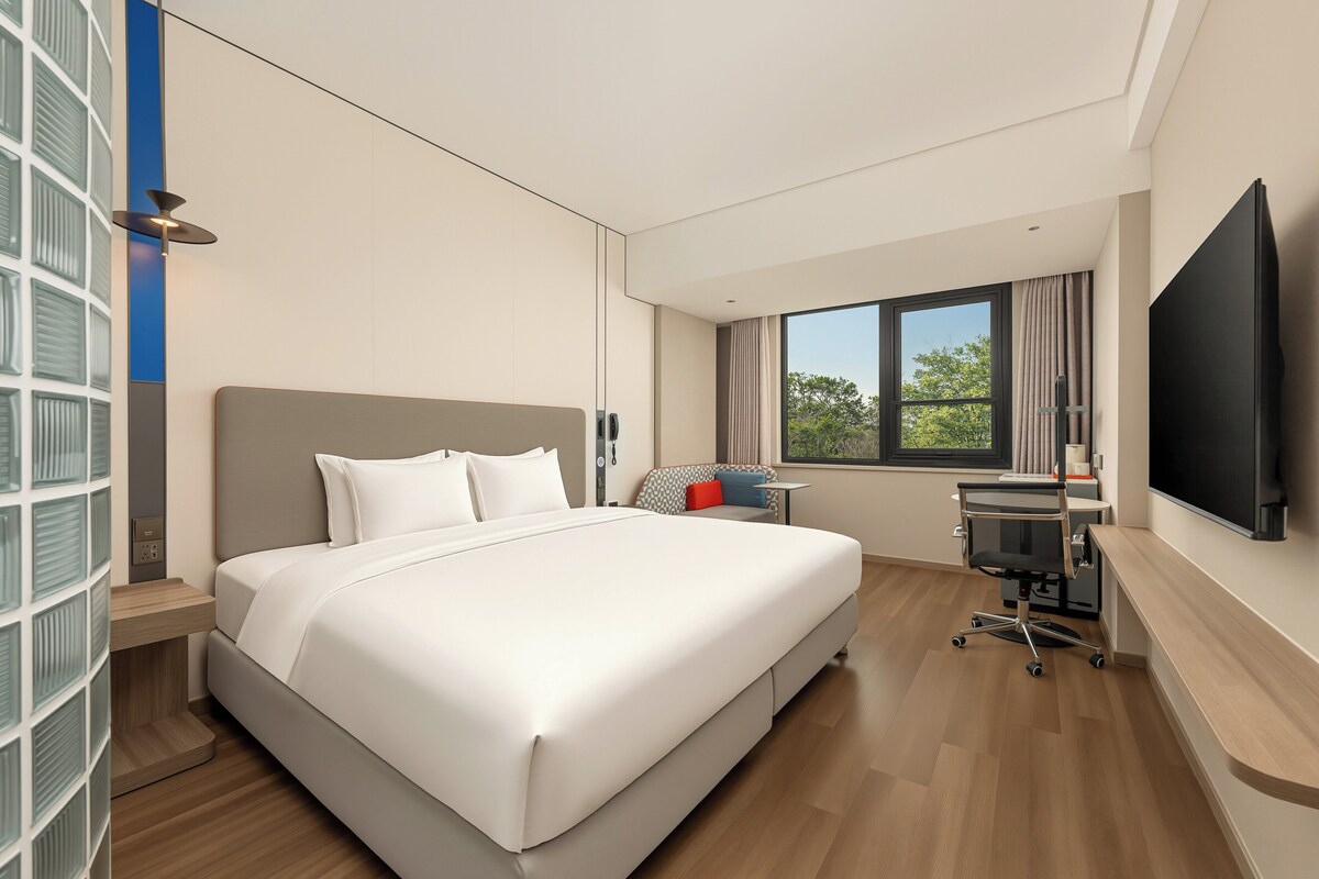 A modern hotel room with a large bed, a desk with a chair, a TV, and a window with a view of trees.