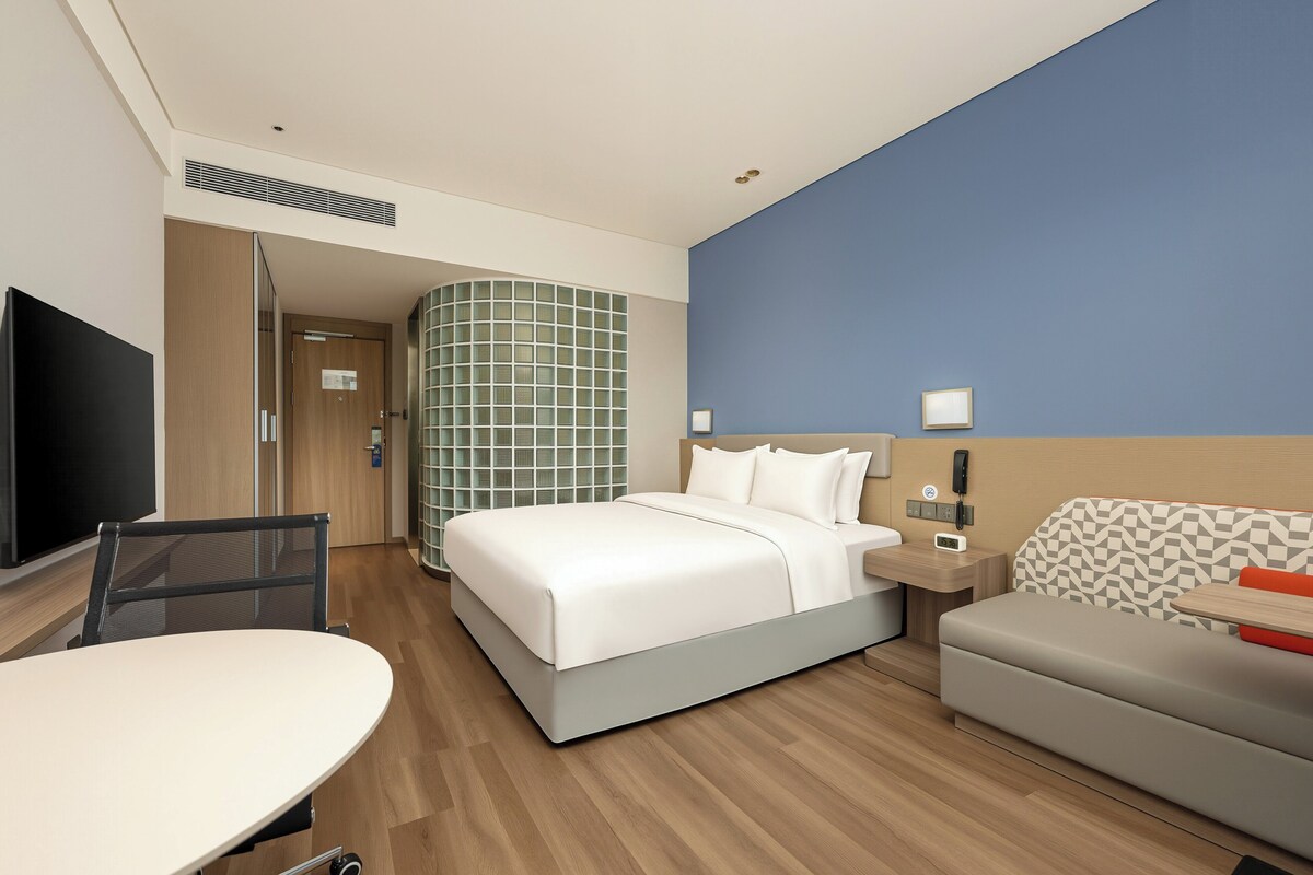 A hotel room with a bed, a desk, a chair, a sofa, a TV, and a bathroom with a shower and a tiled wall.