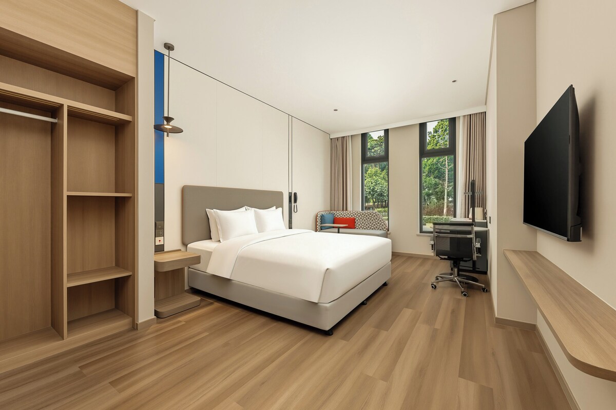A modern hotel room with a bed, a desk, a chair, a TV, and a wardrobe.