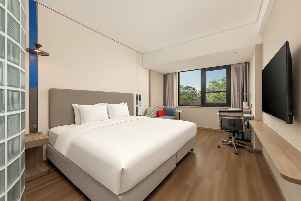 Ein modernes Hotelzimmer mit einem großen Bett, einem Schreibtisch mit Stuhl, einem Fernseher und einem Fenster mit Blick auf Bäume.