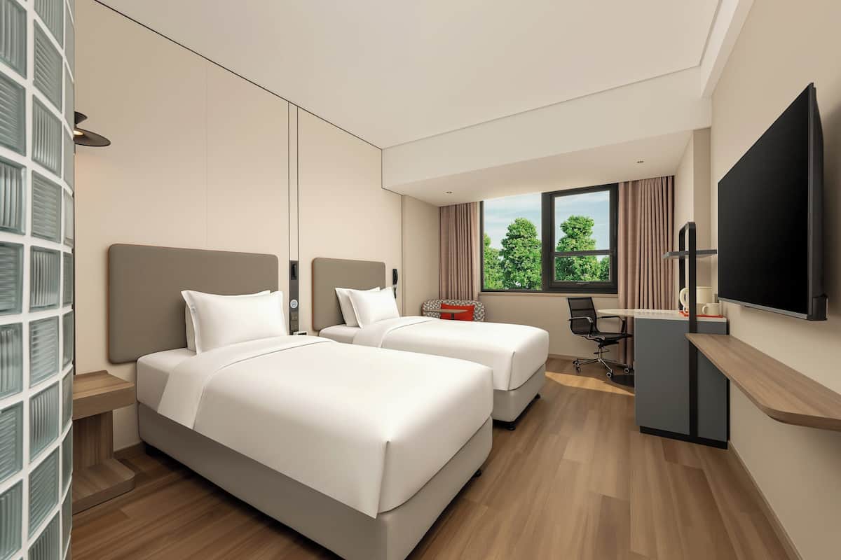 Ein Hotelzimmer mit zwei Betten, einem Schreibtisch, einem Stuhl, einem Fernseher und einem Fenster mit Blick auf Bäume.