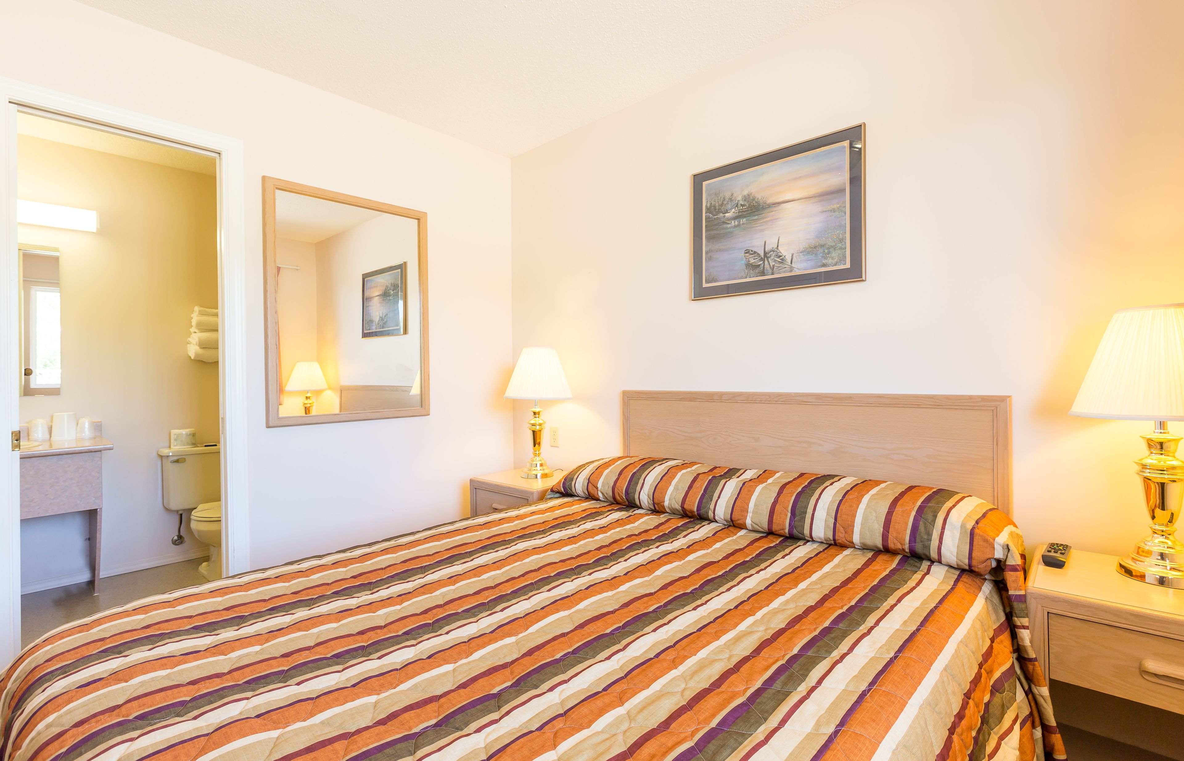 A hotel room with a large bed, a bedside table with a lamp, a nightstand, a mirror, and a painting on the wall.