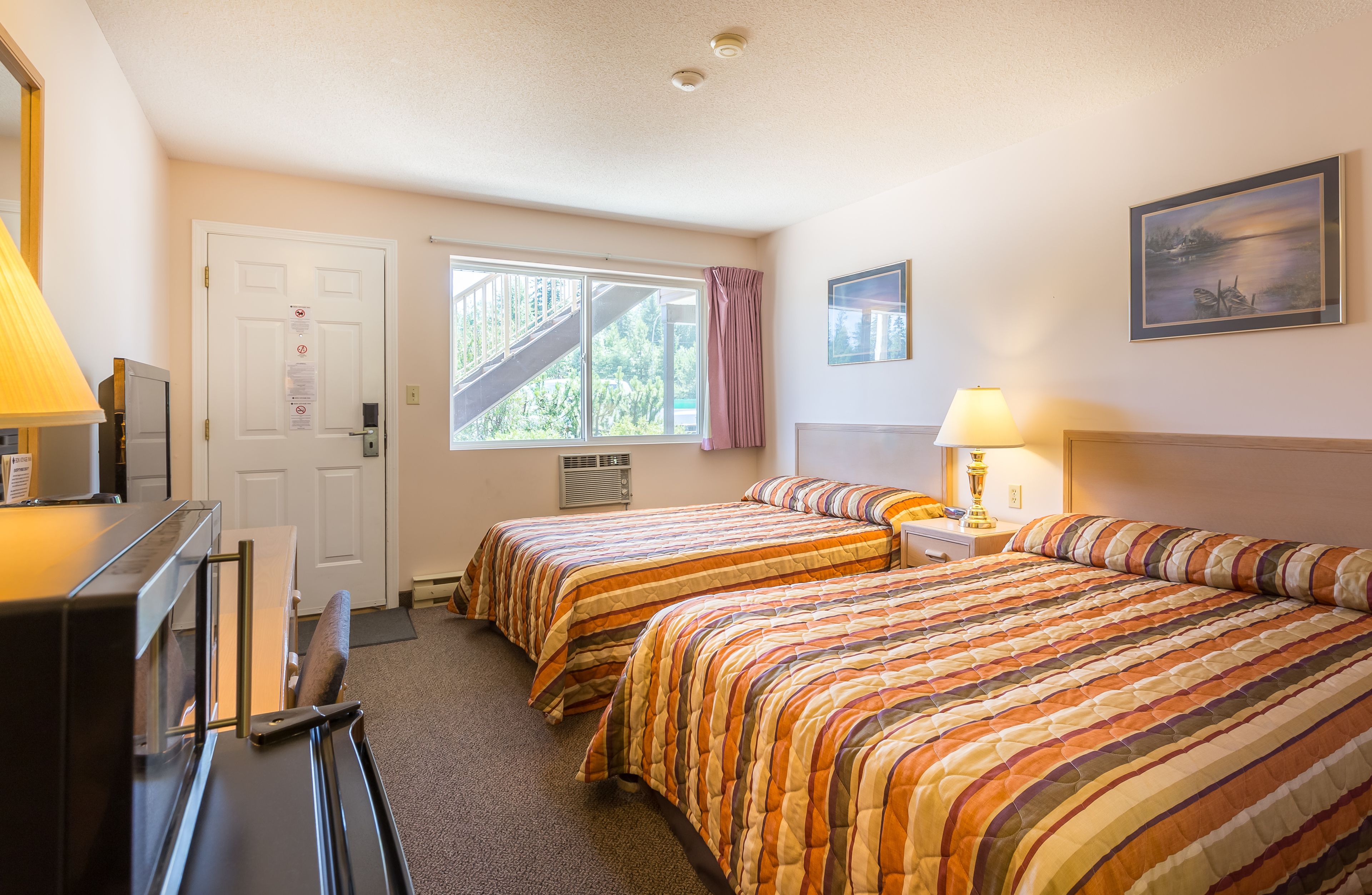 A hotel room with two beds, a desk, a chair, a TV, a window with a view of a staircase, and a painting on the wall.