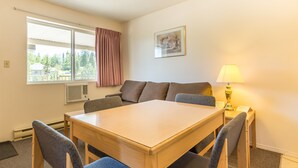 Una habitación con una mesa de madera, cuatro sillas, un ventanal con cortinas, una lámpara de pared y un cuadro enmarcado.