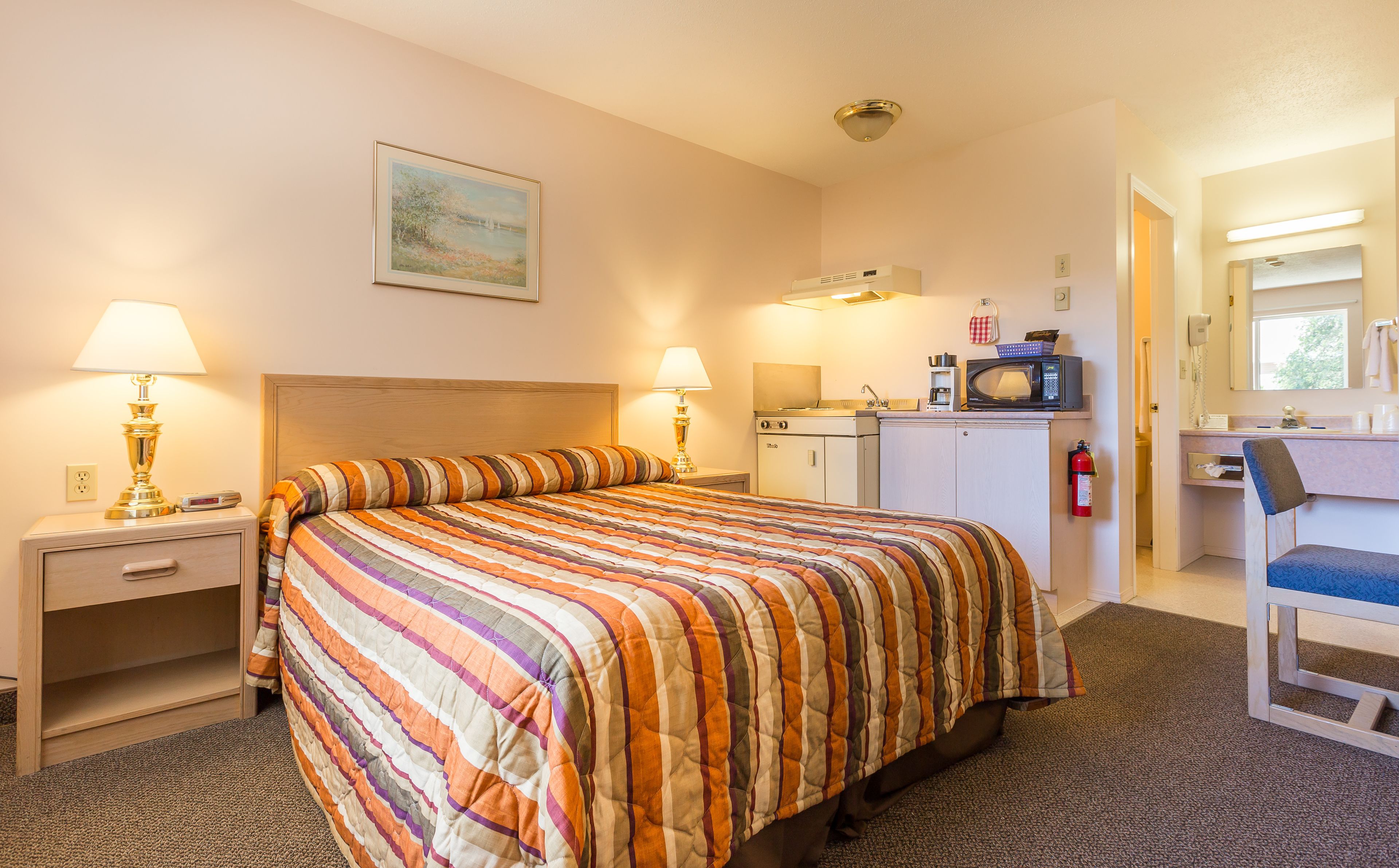 A hotel room with a bed, two bedside tables with lamps, a small kitchenette, a mirror, and a painting on the wall.