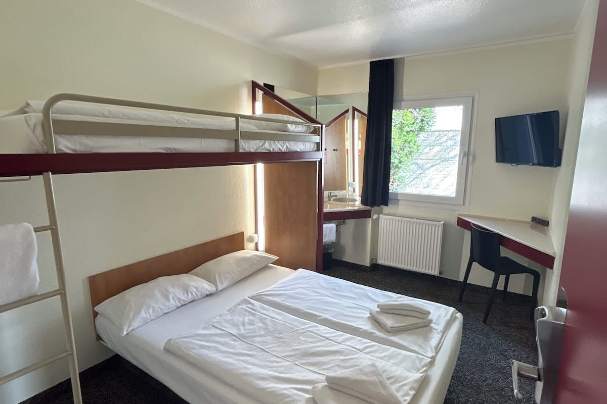 Quarto de hotel com beliches, uma escrivaninha, uma cadeira, uma TV e uma janela com vista.