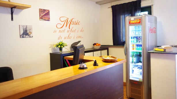 Zona de recepción de un hotel con mostrador de madera, refrigerador Coca-Cola, pared con cita sobre música y ventana con cortinas.