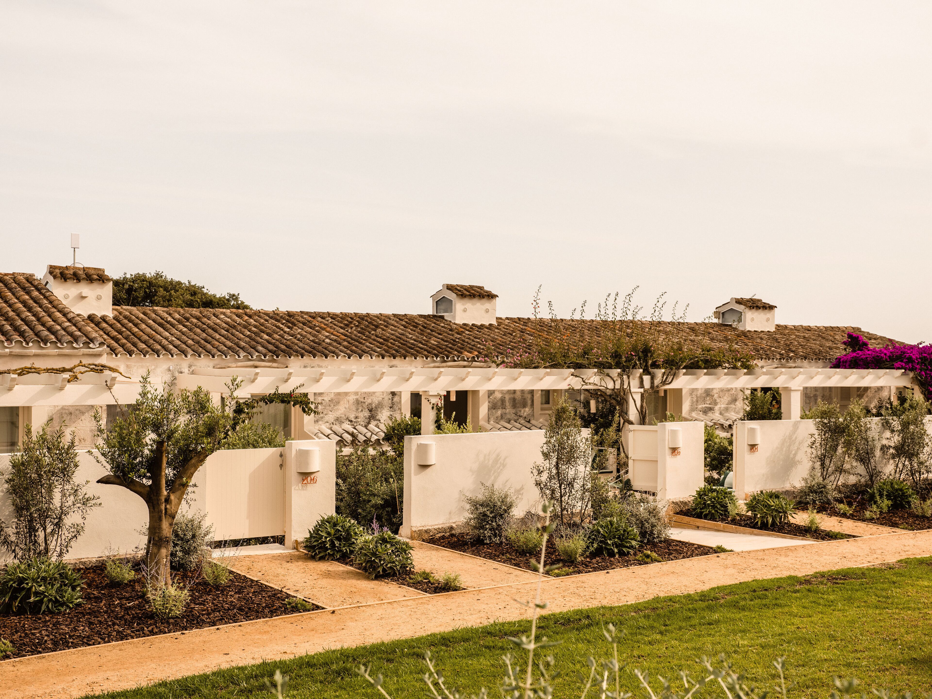 A row of white buildings with tiled roofs, surrounded by a garden with various plants and a gravel path.
