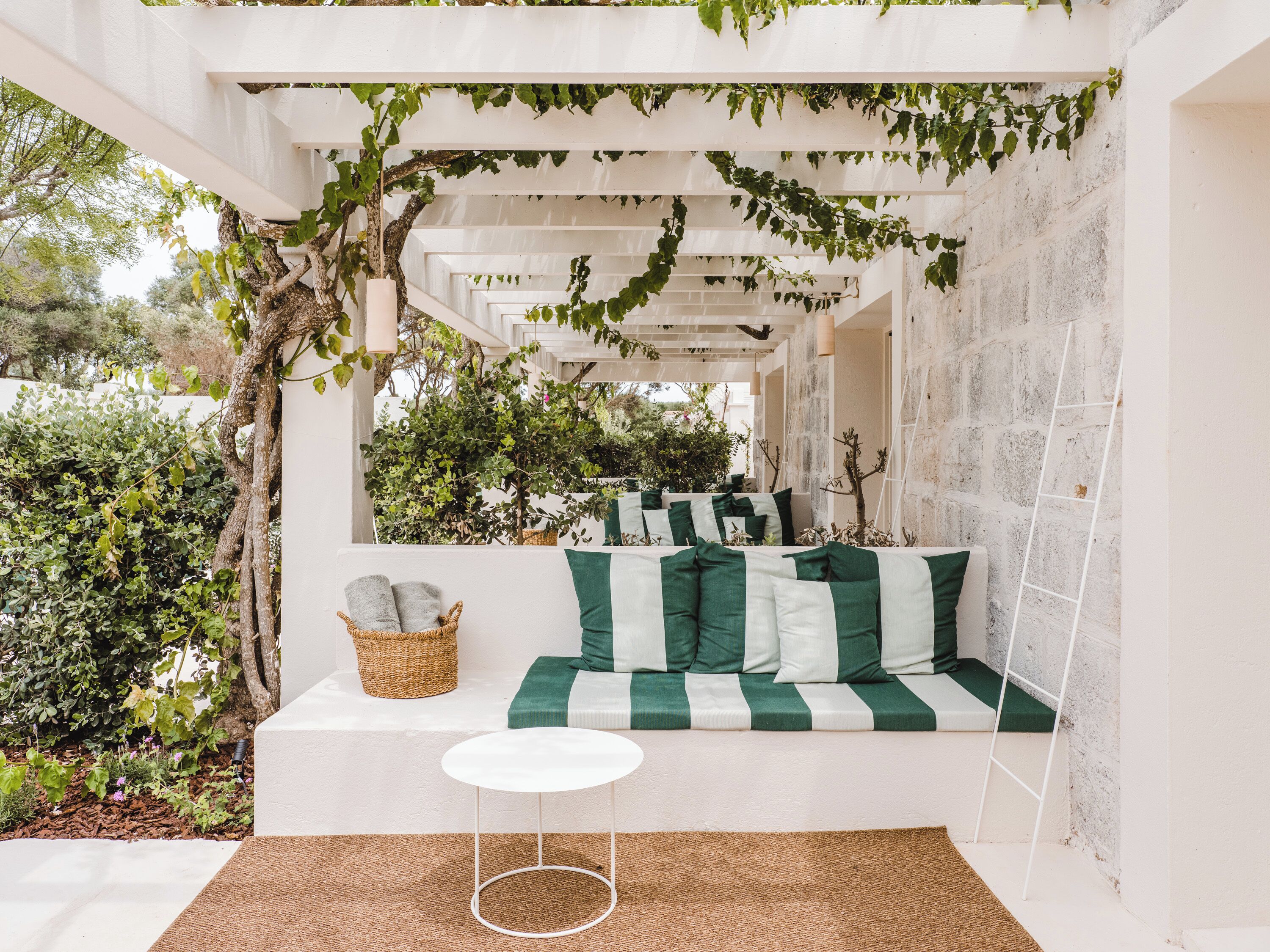 Ein gemütlicher Außenbereich mit einer weißen Couch und Kissen, einem kleinen runden Tisch und einer mit Grünpflanzen bewachsenen Pergola.