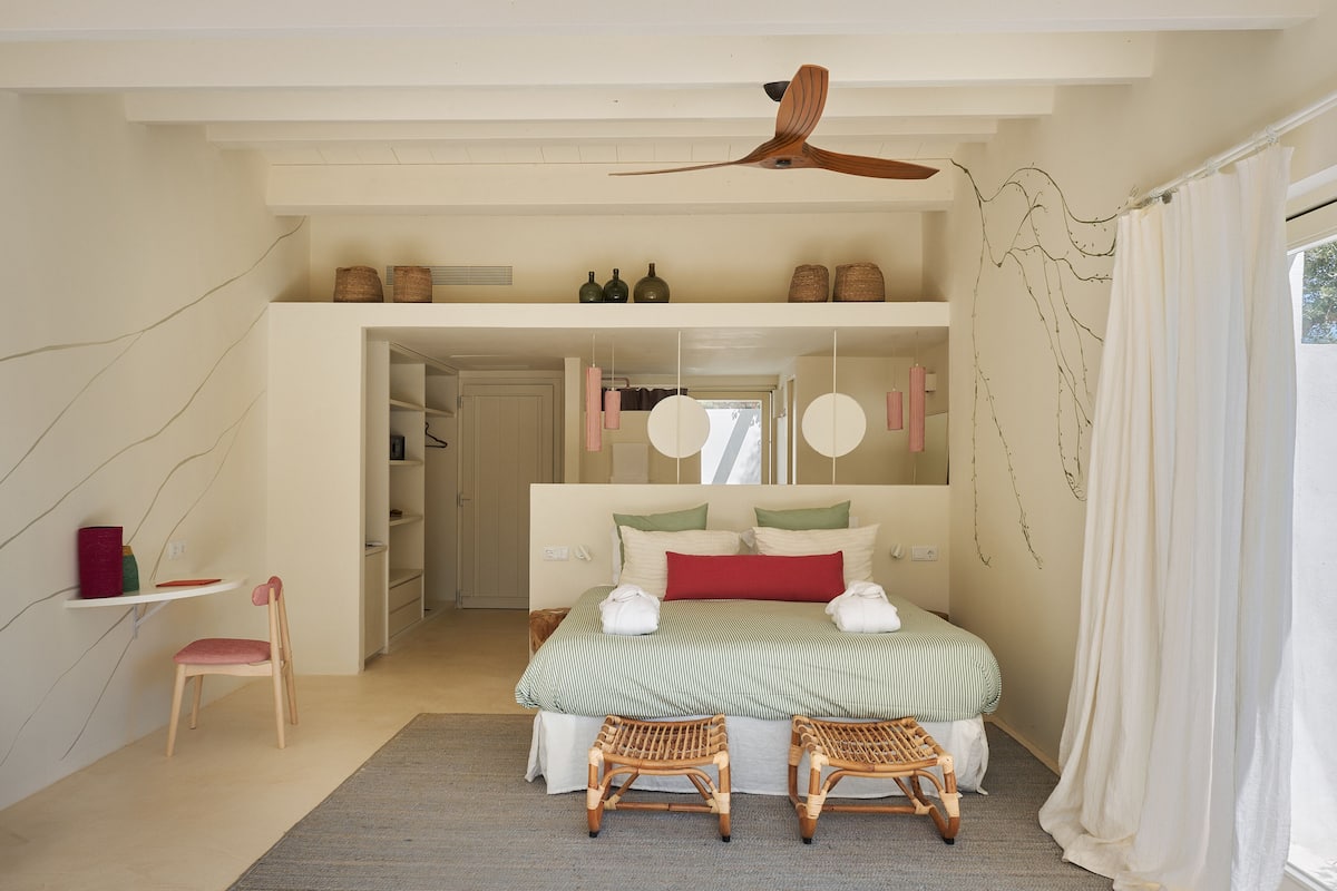 A bedroom with a bed, two chairs, a ceiling fan, a window with curtains, and a hallway leading to a bathroom.