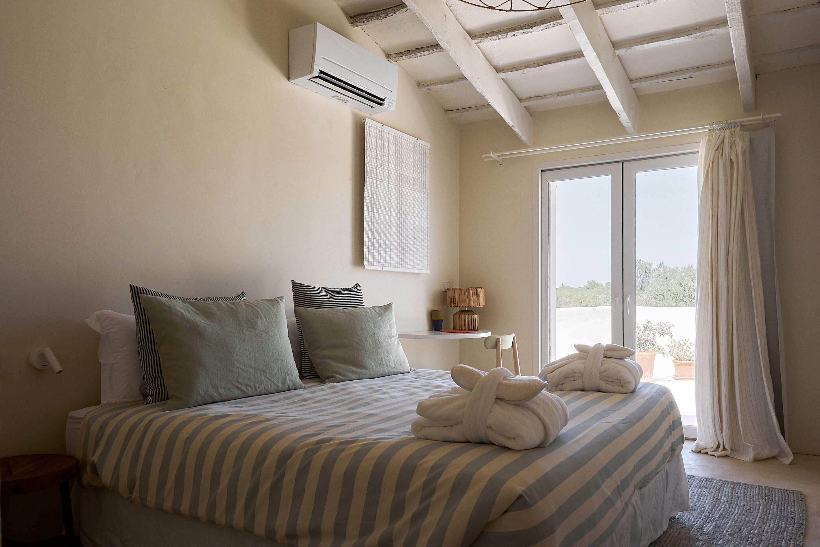 A bedroom with a bed, pillows, a bedside table, an air conditioner, a window with curtains, and a view of the outdoors.