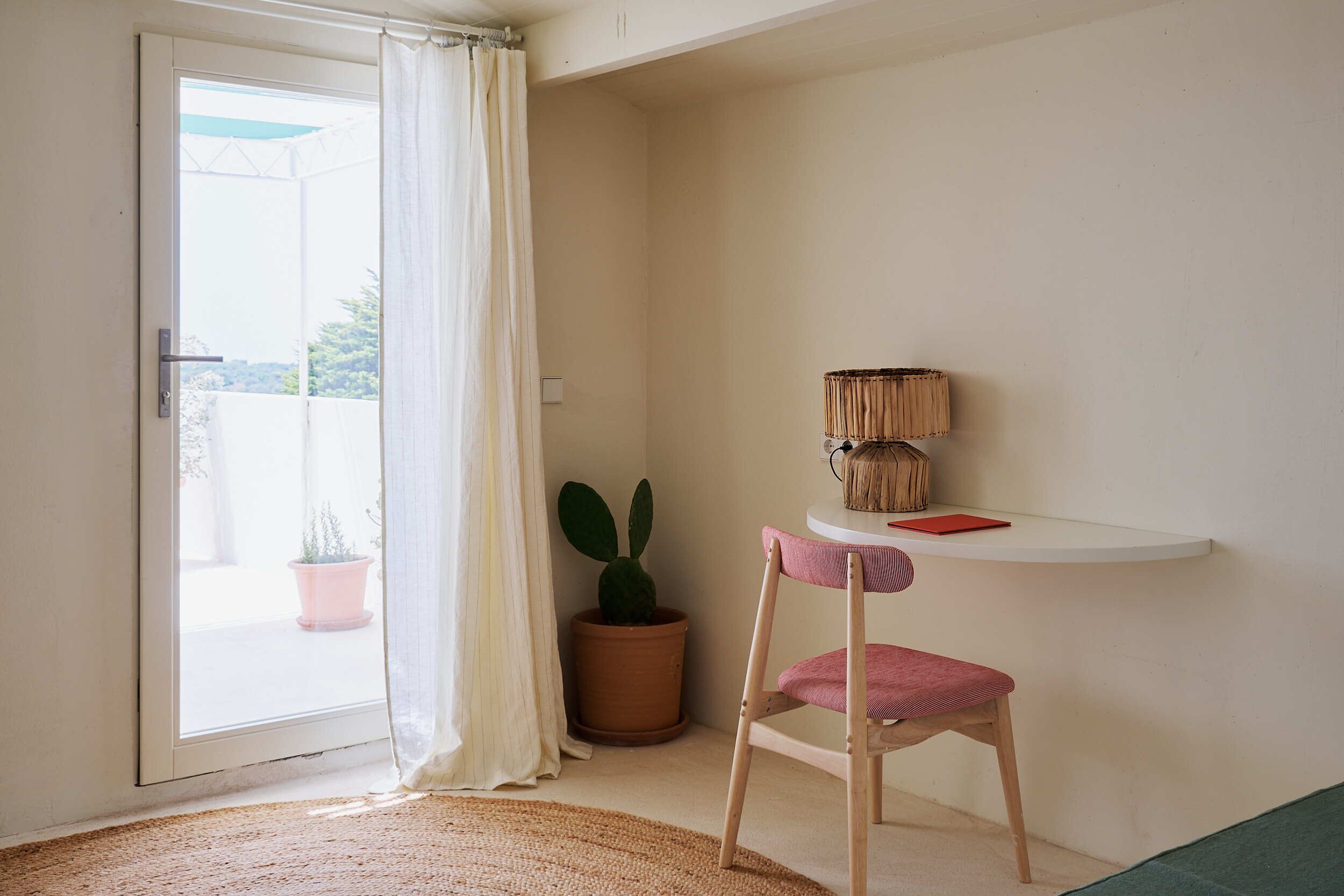 Une pièce avec une porte-fenêtre coulissante, un cactus en pot, une chaise en bois et une petite étagère blanche sur laquelle repose un livre rouge.