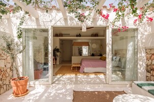 Un dormitorio con una cama, una mesita de noche y una ventana. Hay una planta en maceta y un ventilador de techo.