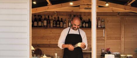 A chef preparing food in a rustic kitchen with wooden walls and shelves of bottles.