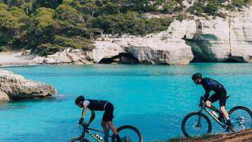 Dos ciclistas en un camino costero con acantilados rocosos y un mar azul y despejado al fondo.