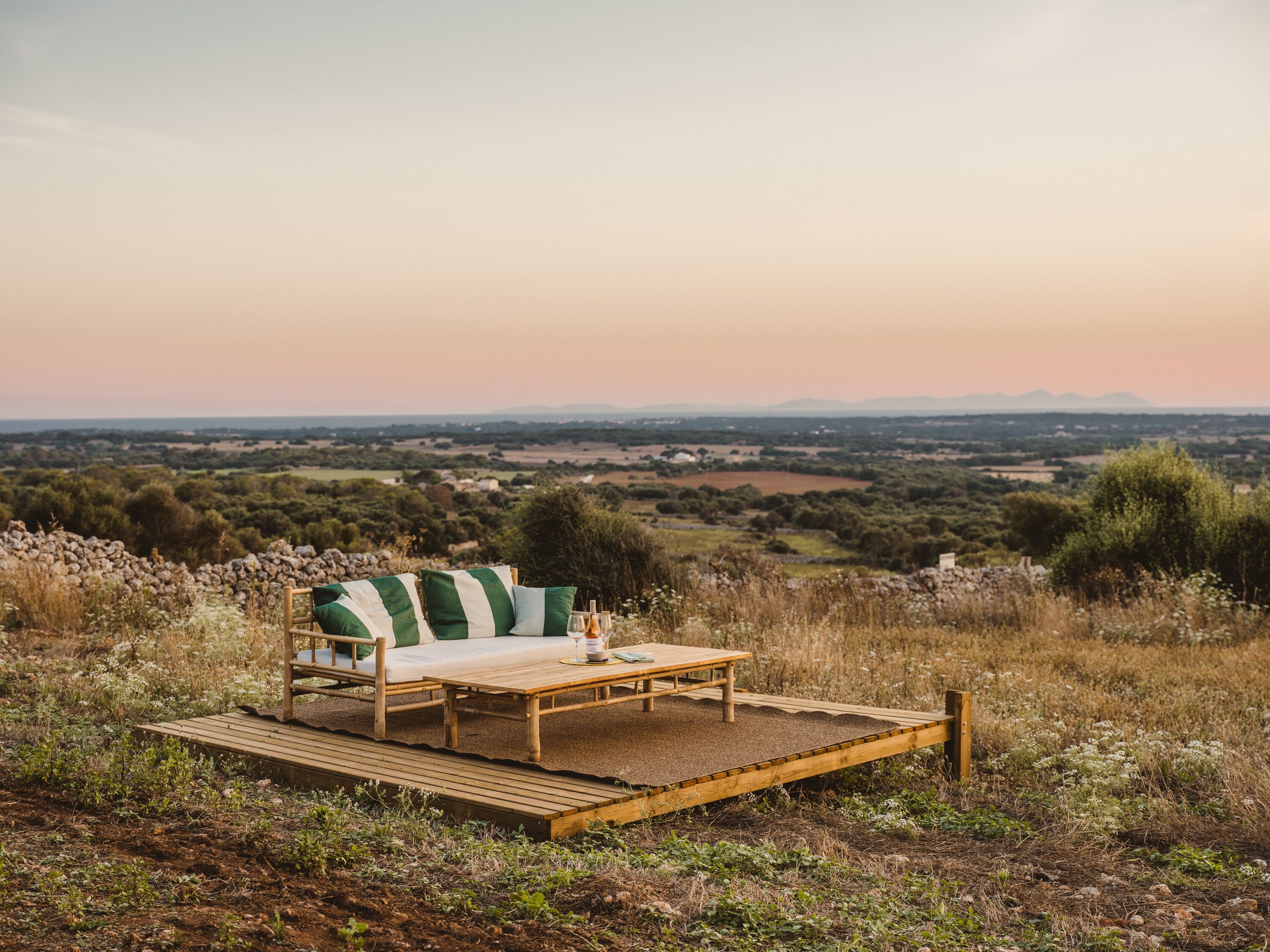 A wooden deck with a sofa and table set up for outdoor dining, overlooking a scenic landscape with distant hills and fields.