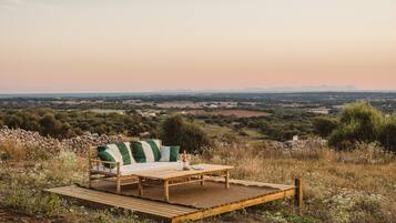 A wooden deck with a sofa and table set up for outdoor dining, overlooking a scenic landscape with distant hills and fields.