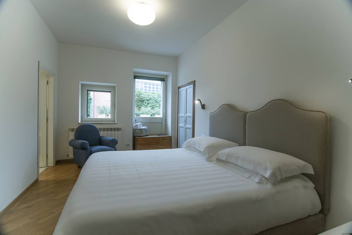A bedroom with a large bed, a chair, two windows, and a door.