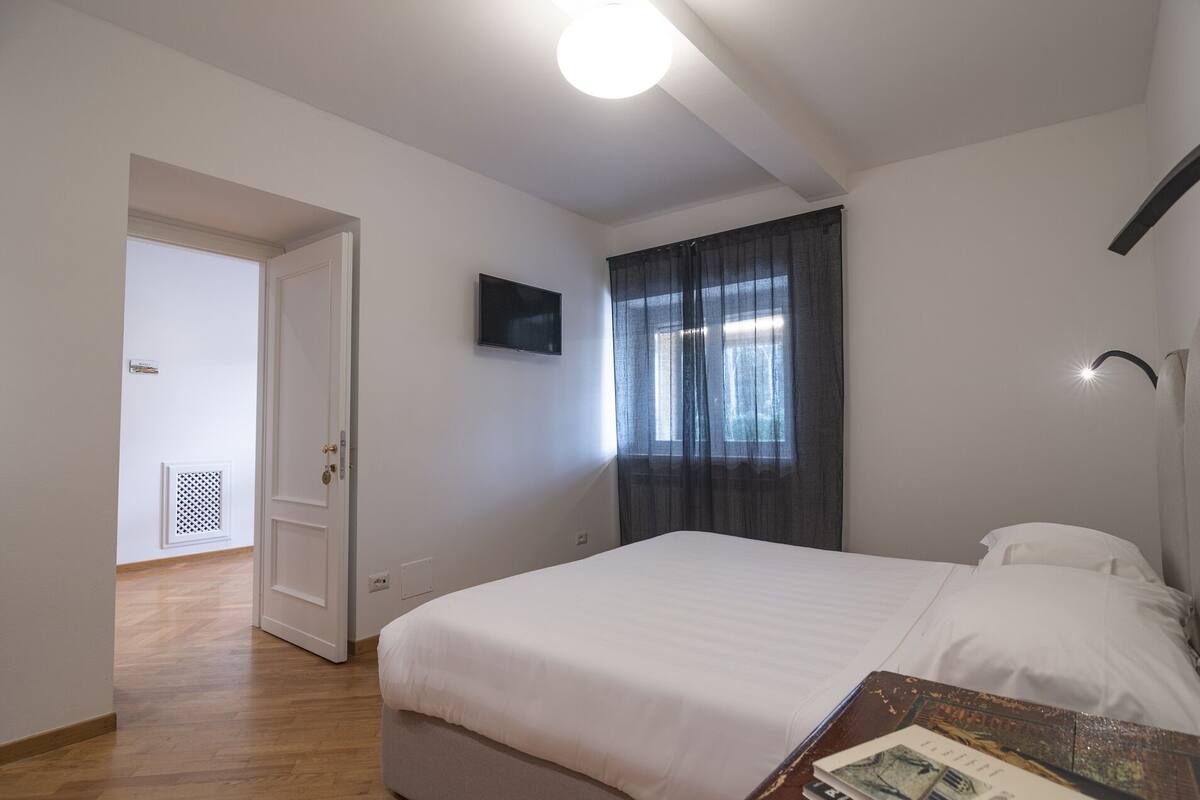 A bedroom with a bed, a flat-screen TV mounted on the wall, a window with curtains, and a door leading to another room.