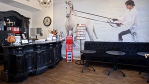 A reception area with a black desk, a large clock, a chandelier, a sofa, and a wall mural of a person leading a horse. - Hotel Iron Horse (Amsterdam)